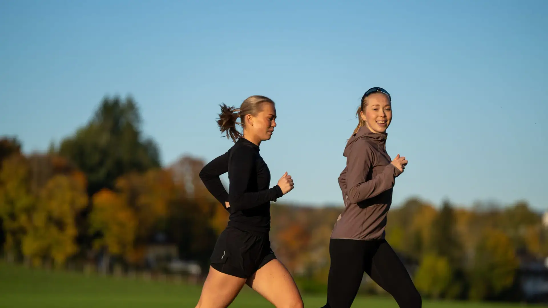 To kvinner løper sammen ute på en gressbane i sollys, omgitt av høstfarger.